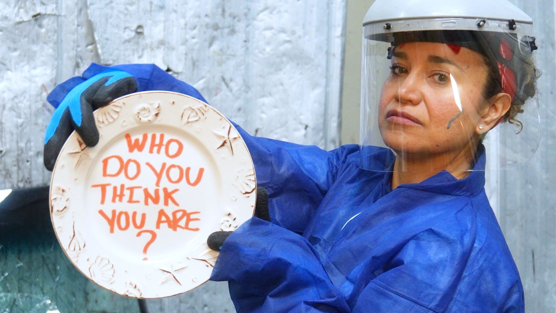 Veronica in blue protective coveralls and a face shield holds a plate reading “Who do you think you are?” in the rage room.