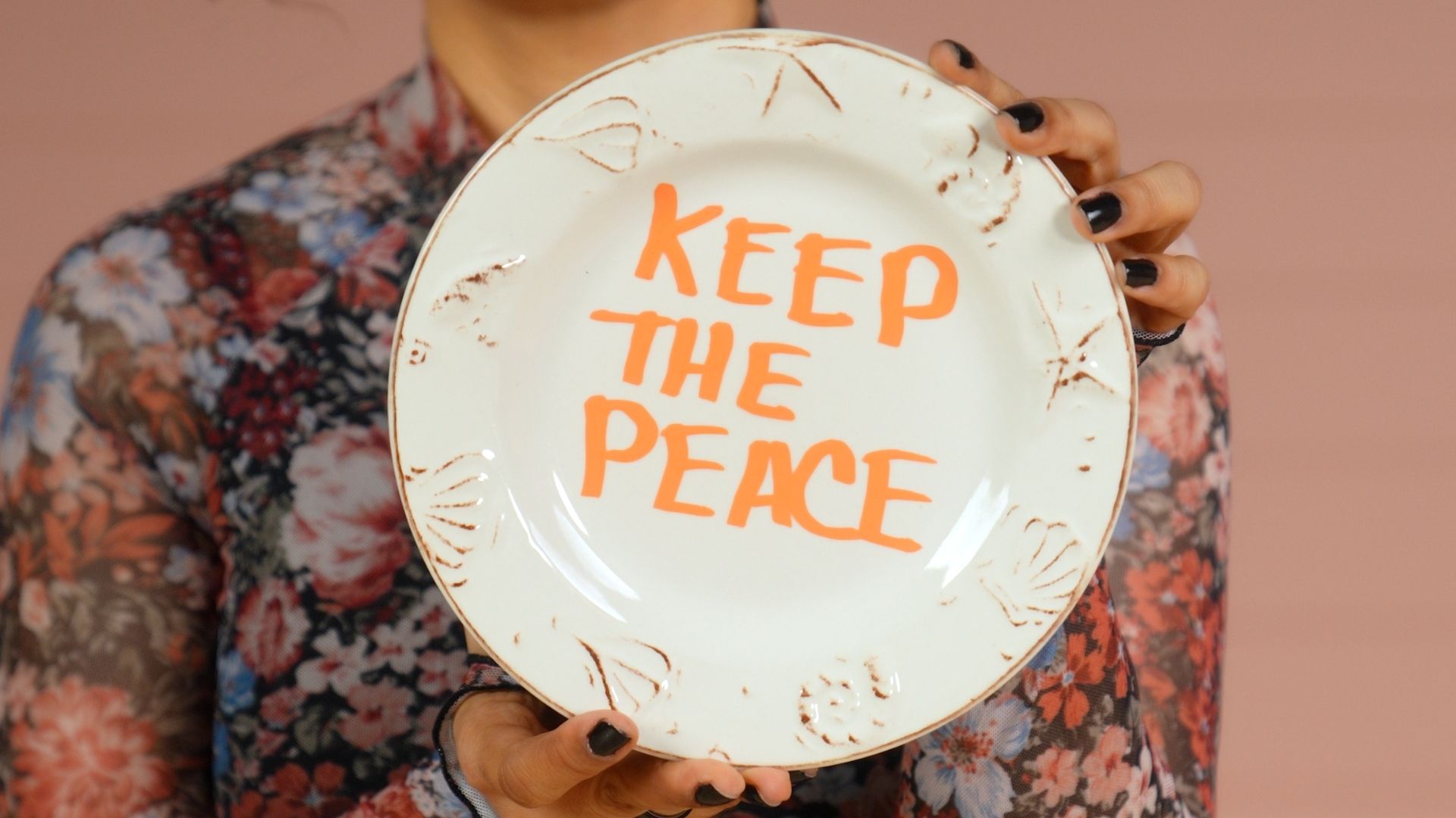 Veronica holds a floral plate close to the camera with “Keep the Peace” written in orange.
